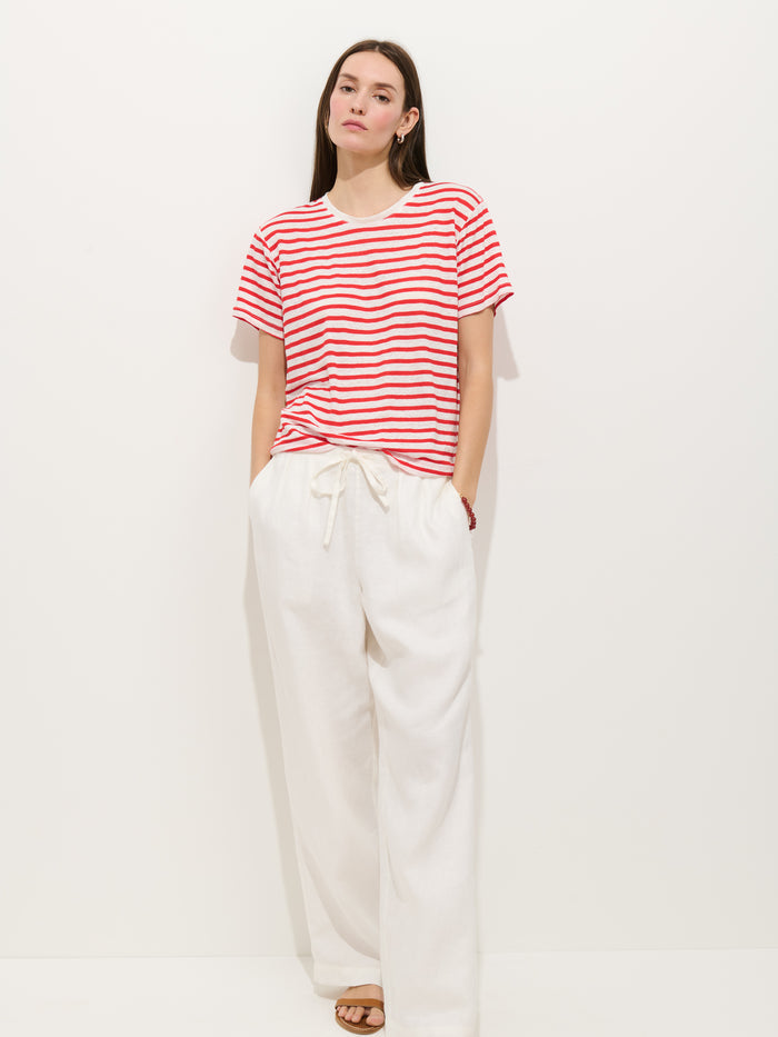A woman with long straight hair and a neutral expression stands against a white background wearing the Garcon Striped Tee in Linen Cotton, loose white drawstring pants, and sandals.