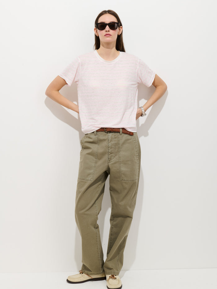 A person stands against a white background wearing black sunglasses, the Garcon Striped Tee in Linen Cotton, loose olive green pants with a brown belt, beige shoes, and poses with hands on hips for timeless style.