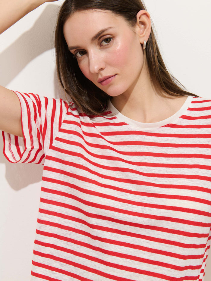 A woman with long brown hair wears the Garcon Striped Tee In Linen Cotton, featuring red and white stripes, as she stands against a white wall with one arm raised and looks at the camera with a neutral expression.