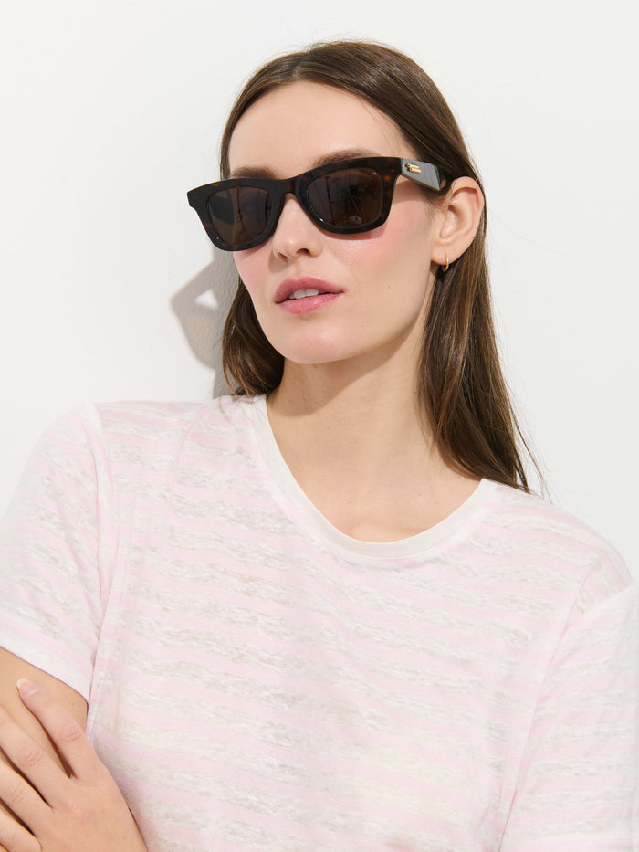 A woman with long brown hair wears black sunglasses and the Garcon Striped Tee In Linen Cotton in a classic fit, standing against a white background with her arms crossed—perfect for effortless style year round.