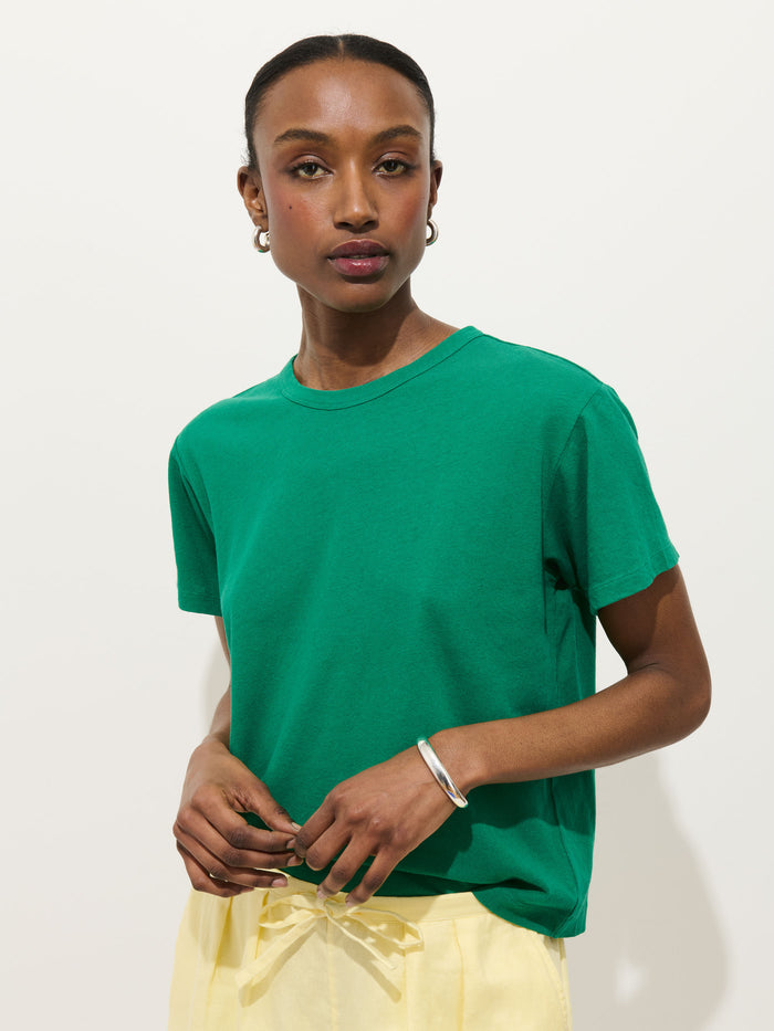 A woman with dark hair pulled back wears the Garcon Tee In Linen Cotton in green and light yellow pants, styled with hoop earrings and a silver bracelet. She stands against a plain white background, looking confidently at the camera.