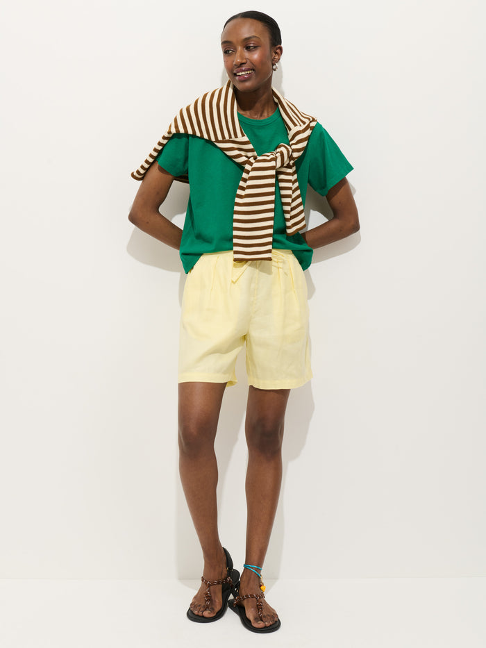 A person stands smiling against a plain white background, wearing the Garcon Tee In Linen Cotton, pale yellow shorts, black sandals, and a brown-and-white striped sweater draped over their shoulders.