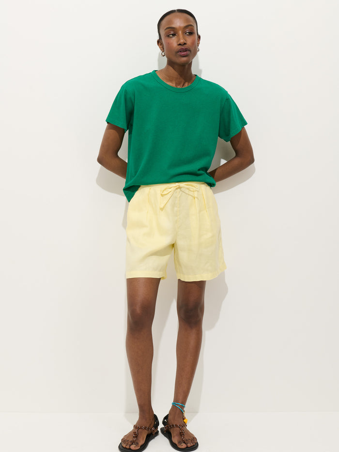 A person stands against a white background wearing the Garcon Tee in Linen Cotton (green), light yellow shorts, brown sandals, and an anklet. Hands are in pockets as they look at the camera.