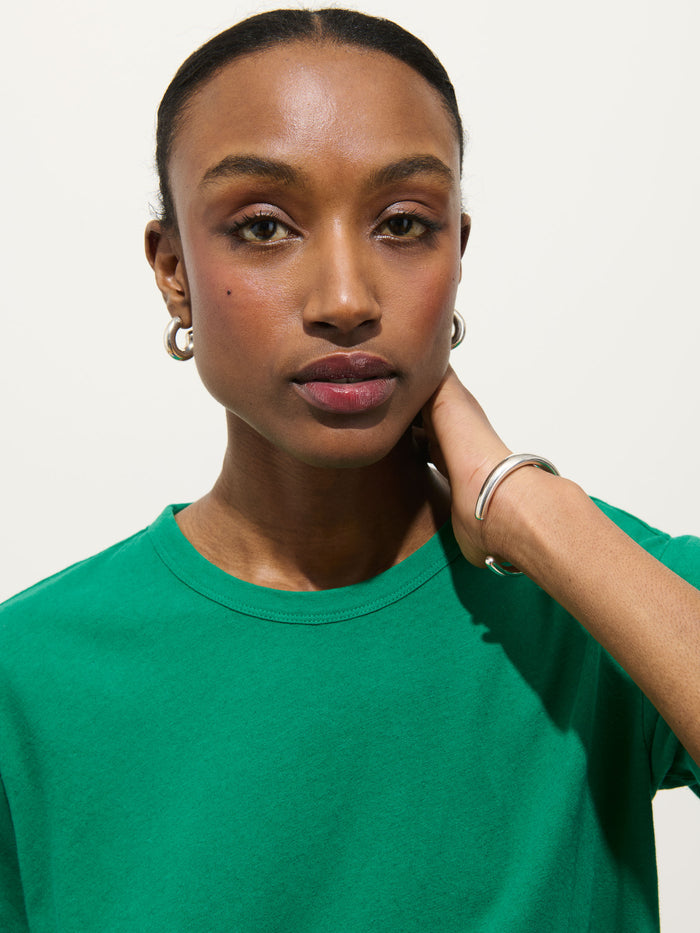 A woman in the Garcon Tee In Linen Cotton, paired with silver hoop earrings and a silver bracelet, confidently looks at the camera, touching her neck against a plain light background.