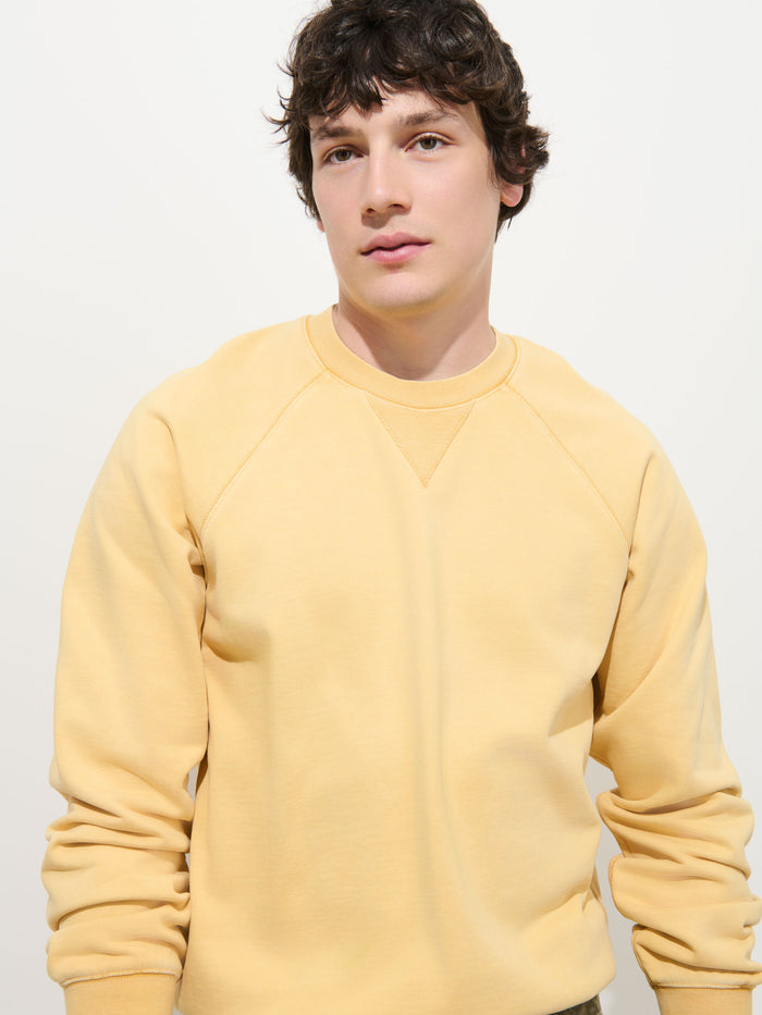 A person with short brown hair wearing the Garment Dye Raglan Sweatshirt in Cotton Fleece, featuring a light yellow, vintage-inspired design, stands against a plain white background and looks slightly to the side with a neutral expression.