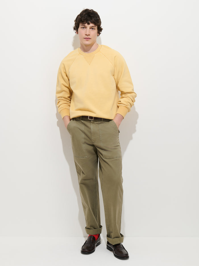 A person stands against a white wall wearing the Garment Dye Raglan Sweatshirt in Cotton Fleece in light yellow, paired with olive green pants and dark loafers. With hands in pockets and a neutral expression, the vintage-inspired sweatshirt lends a subtle retro touch to the outfit.