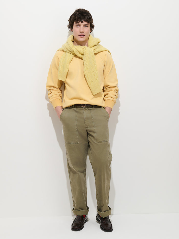 A person stands against a white background, wearing the Garment Dye Raglan Sweatshirt in Cotton Fleece in light yellow, draped with a yellow sweater over their shoulders, paired with olive green pants and dark brown shoes. They have short curly hair, hands in pockets, and look directly at the camera.