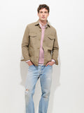A person stands against a plain white background with hands in pockets, wearing the Garment Dyed Chore Jacket in Canvas—a vintage hunting-inspired piece crafted from garment-dyed canvas—layered over a pink sweater, white shirt, and light blue ripped jeans.
