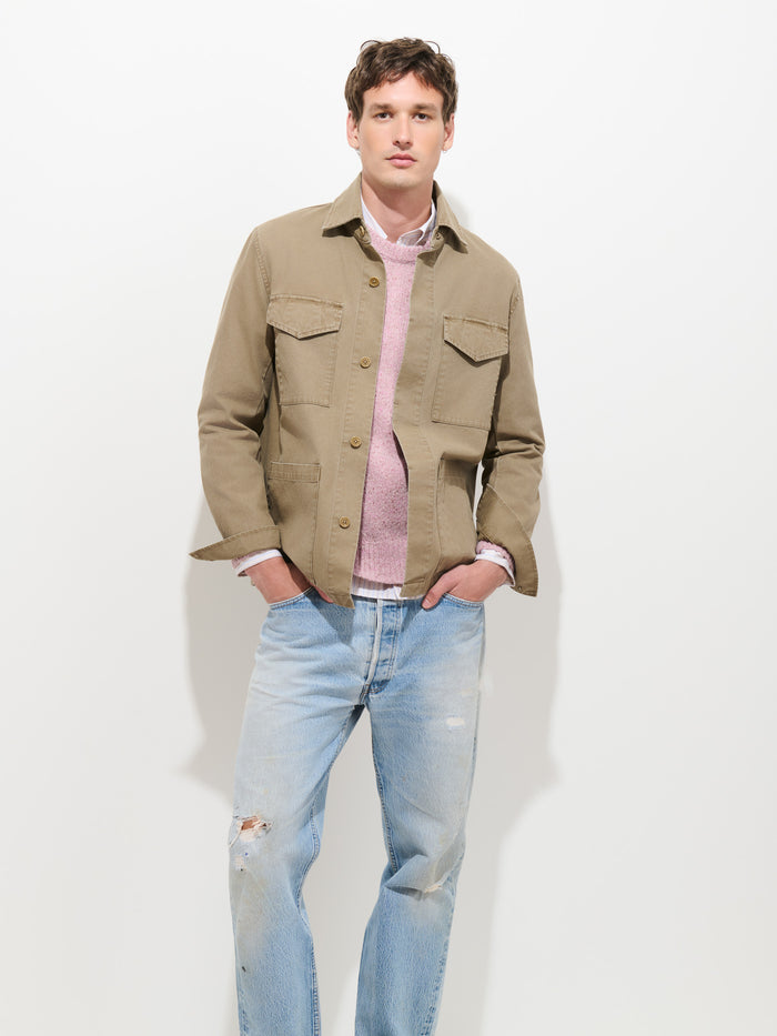 A person stands against a plain white background with hands in pockets, wearing the Garment Dyed Chore Jacket in Canvas—a vintage hunting-inspired piece crafted from garment-dyed canvas—layered over a pink sweater, white shirt, and light blue ripped jeans.