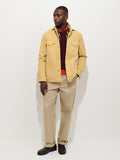 A man stands against a white background wearing the Garment Dyed Chore Jacket in Canvas—a yellow, vintage hunting-inspired jacket—paired with a burgundy sweater, red shirt, beige pants, and dark brown shoes.