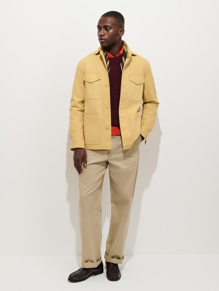 A man stands against a white background wearing the Garment Dyed Chore Jacket in Canvas—a yellow, vintage hunting-inspired jacket—paired with a burgundy sweater, red shirt, beige pants, and dark brown shoes.