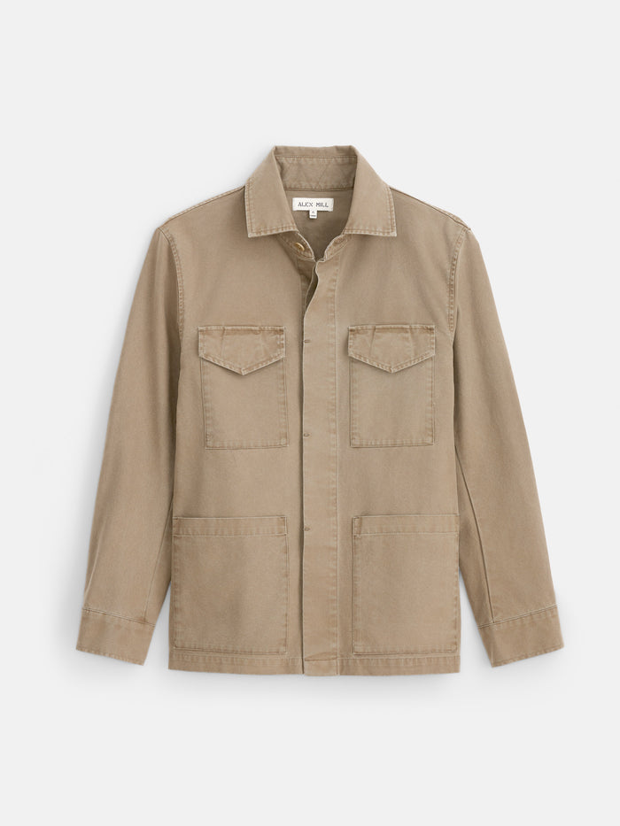 Displayed flat against a white background, the Garment Dyed Chore Jacket in Canvas features a tan hue and long sleeves, crafted from garment-dyed canvas. Designed with a classic collar, two chest flap pockets, and two large lower pockets, this utility jacket draws vintage inspiration from timeless hunting styles while offering practical detailing.