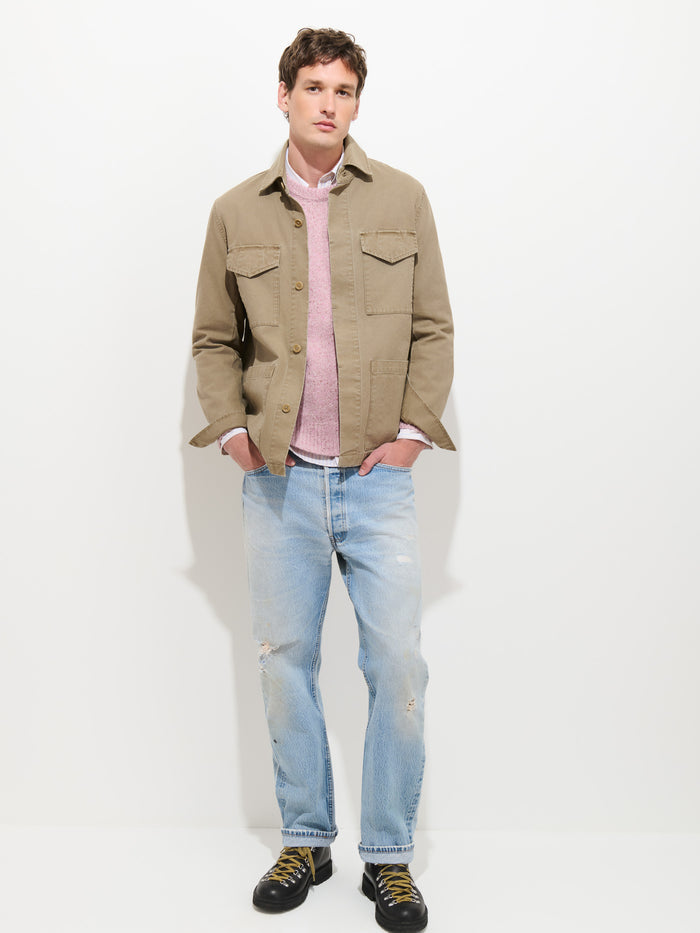 A man stands against a white background wearing the Garment Dyed Chore Jacket in Canvas—a tan, garment-dyed piece—layered over a pink sweater and white shirt, paired with light blue distressed jeans and black sneakers featuring yellow laces. With his hands in his pockets, he blends vintage hunting-inspired style with modern casual elements.