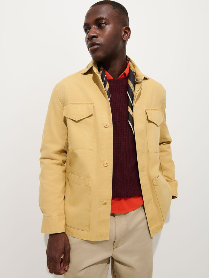 A man wearing the Garment Dyed Chore Jacket in Canvas, styled in a vintage hunting-inspired, light yellow hue over a burgundy sweater, orange shirt, and striped scarf, stands against a plain white background.
