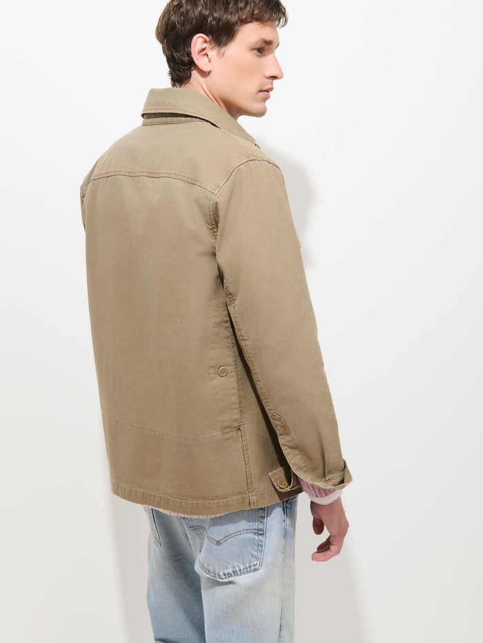 A person wearing the Garment Dyed Chore Jacket in Canvas in tan and light blue jeans is standing and facing away from the camera against a plain white background.