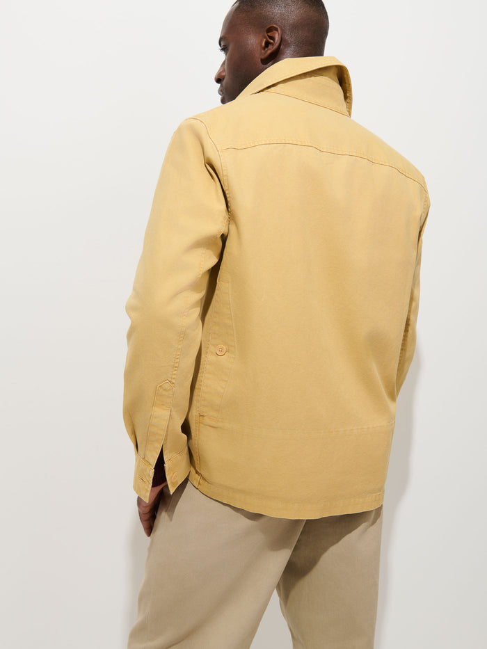A person stands with their back to the camera, wearing the Garment Dyed Chore Jacket in Canvas—a light beige, vintage hunting-inspired jacket with the collar turned up—paired with matching beige pants. The look is crafted from garment-dyed canvas and is set against a plain white background.