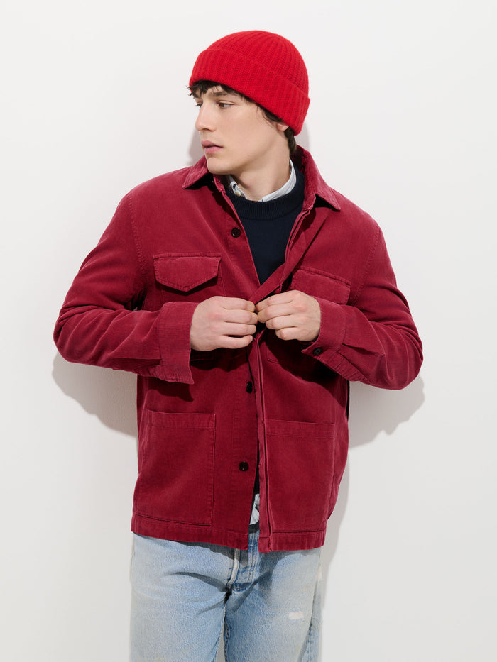 A person wearing a red beanie, a Garment Dyed Chore Jacket in Corduroy with corozo buttons, a black sweater, and light blue jeans stands against a white background, looking to the side and adjusting their jacket buttons.