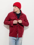 A person wearing a red beanie, a Garment Dyed Chore Jacket in Corduroy with corozo buttons, a black sweater, and light blue jeans stands against a white background, looking to the side and adjusting their jacket buttons.