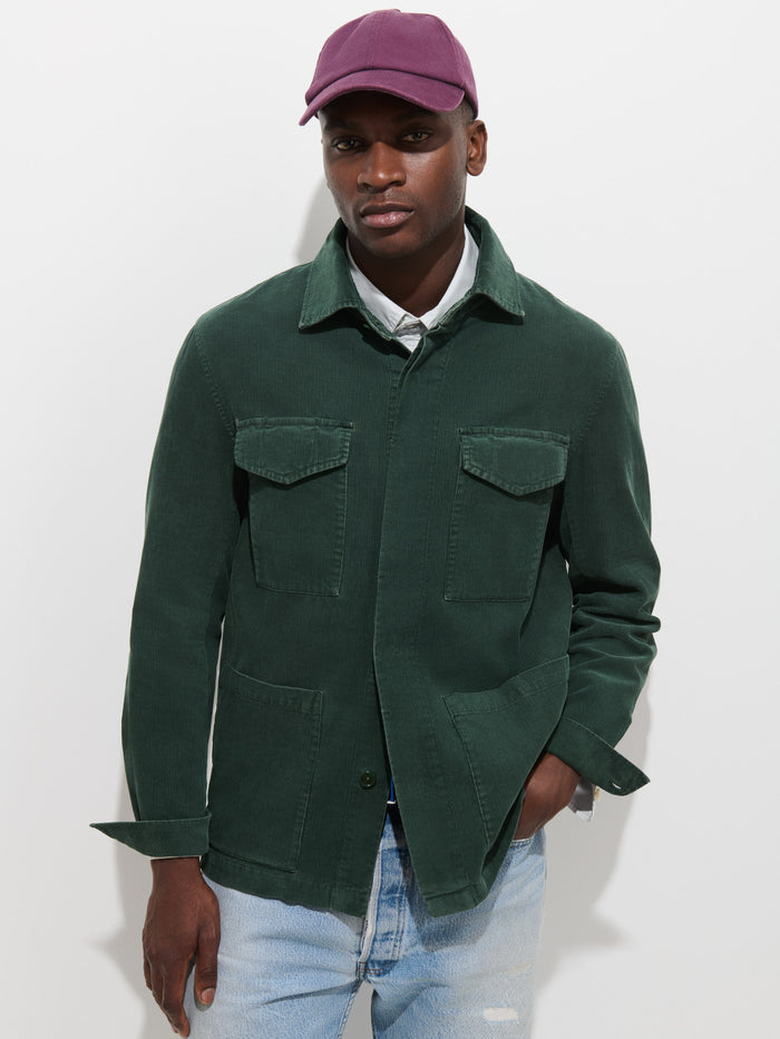 A person wearing a purple cap, the Garment Dyed Chore Jacket in Corduroy featuring front pockets and corozo buttons in dark green, and light blue ripped jeans stands against a plain white background.