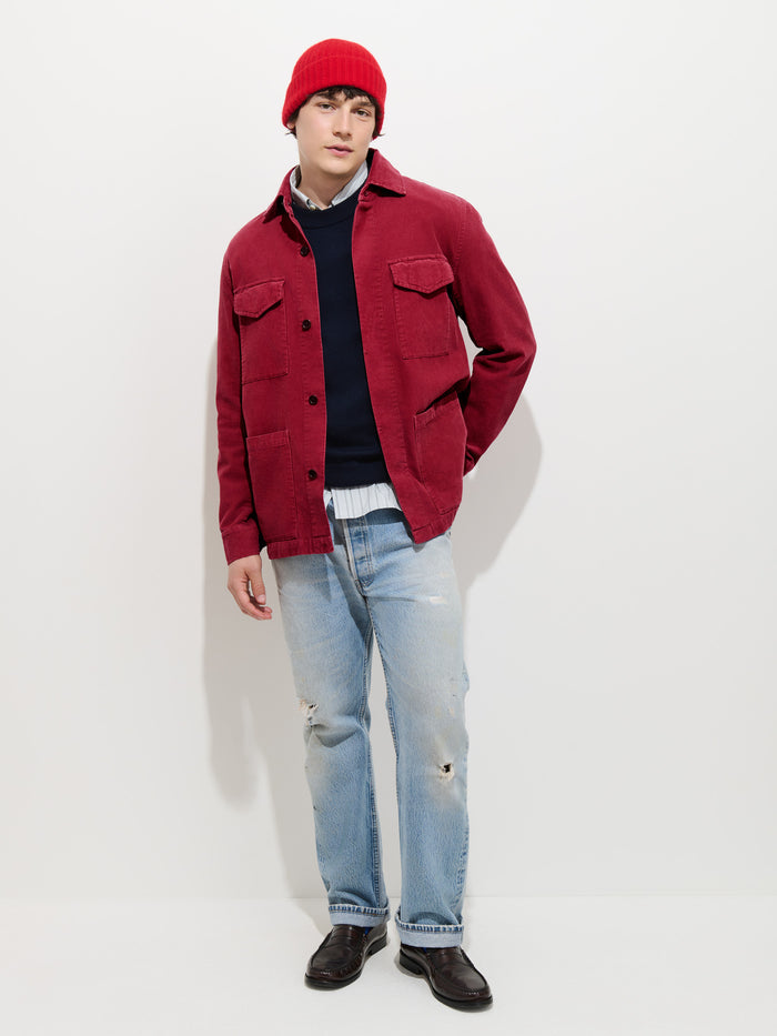 A person wearing a red beanie, the Garment Dyed Chore Jacket in Corduroy with corozo buttons, a black sweater, light blue ripped jeans, and black shoes stands against a plain white background.