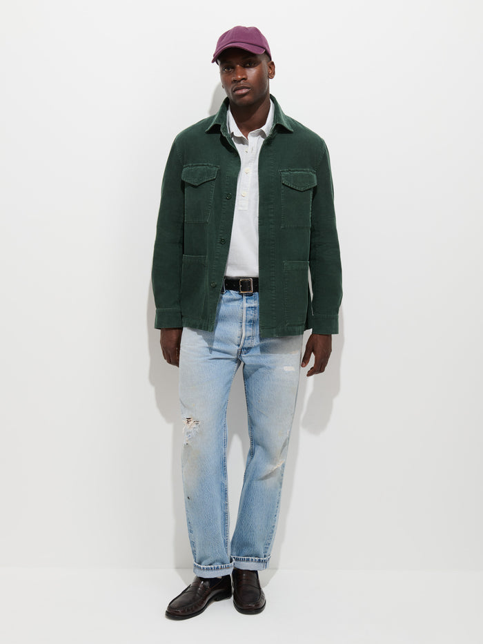 A man stands against a plain white background wearing a maroon cap, the Garment Dyed Chore Jacket in Corduroy with corozo buttons, a white shirt, light blue ripped jeans, and dark shoes.