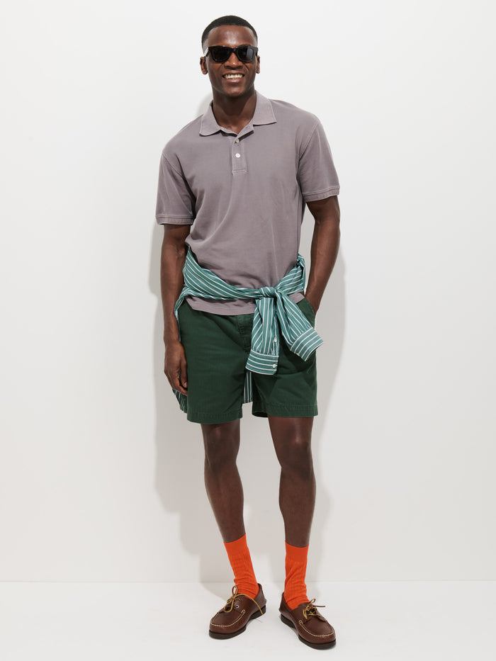 A man smiles against a white background, wearing a Garment Dyed Polo In Pique Cotton in gray, green shorts, orange socks, brown loafers, dark sunglasses, and a blue-green striped shirt tied around his waist.