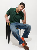 A man in a Garment Dyed Polo In Pique Cotton, blue jeans, and orange socks sits on a black chair, leaning forward with one hand on his head and the other on the chair, looking thoughtfully at the camera against a plain white background.