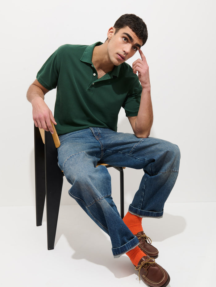 A man in a Garment Dyed Polo In Pique Cotton, blue jeans, and orange socks sits on a black chair, leaning forward with one hand on his head and the other on the chair, looking thoughtfully at the camera against a plain white background.