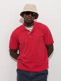 A person wears the Garment Dyed Polo In Pique Cotton in red over a striped shirt, navy pants, beige bucket hat, and black sunglasses while standing against a white background with hands behind their back.