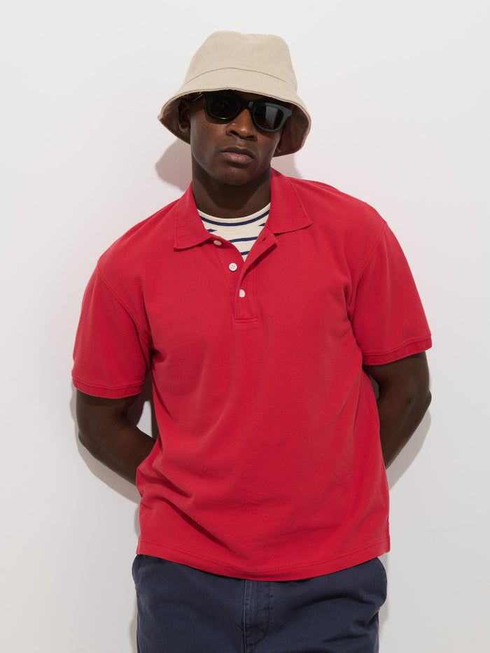 A person wears the Garment Dyed Polo In Pique Cotton in red over a striped shirt, navy pants, beige bucket hat, and black sunglasses while standing against a white background with hands behind their back.
