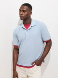 A man stands against a white background wearing the Garment Dyed Polo in Pique Cotton layered over a red shirt with white pants. He looks to the side, one hand in his pocket, maintaining a neutral expression.