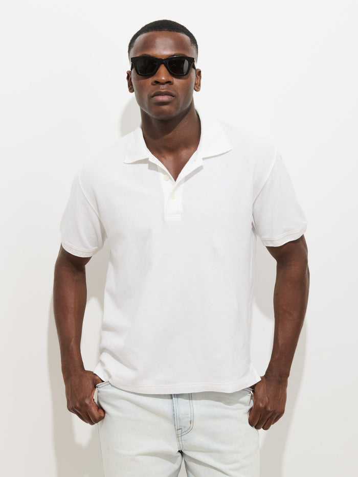 A man wearing black sunglasses, a white Garment Dyed Polo In Pique Cotton, and light blue jeans stands against a plain white background with his hands in his pockets.
