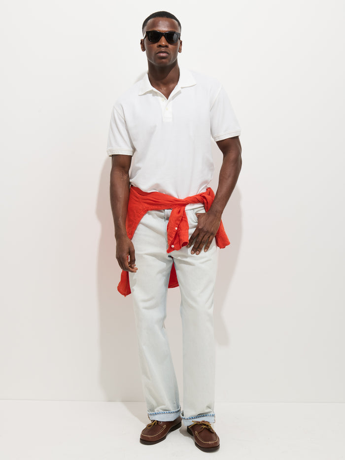 A man stands against a white background wearing sunglasses, a Garment Dyed Polo In Pique Cotton, light blue jeans, brown shoes, and a red sweatshirt tied around his waist.