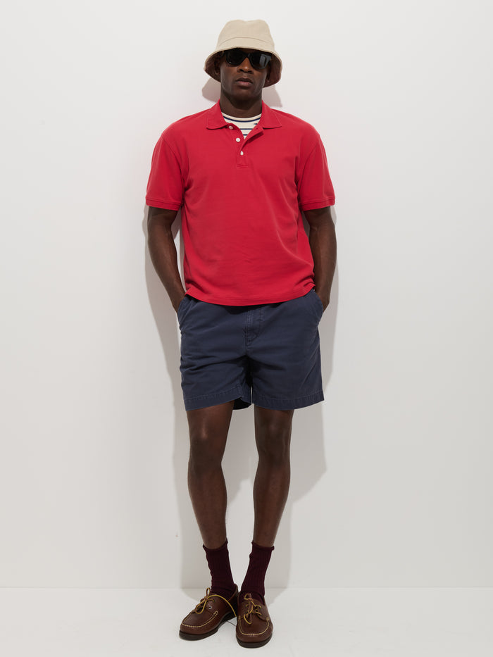 A man stands against a white background wearing a Garment Dyed Polo In Pique Cotton (red), navy blue shorts, maroon socks, brown boat shoes, a beige bucket hat, and black sunglasses, with his hands in his pockets.