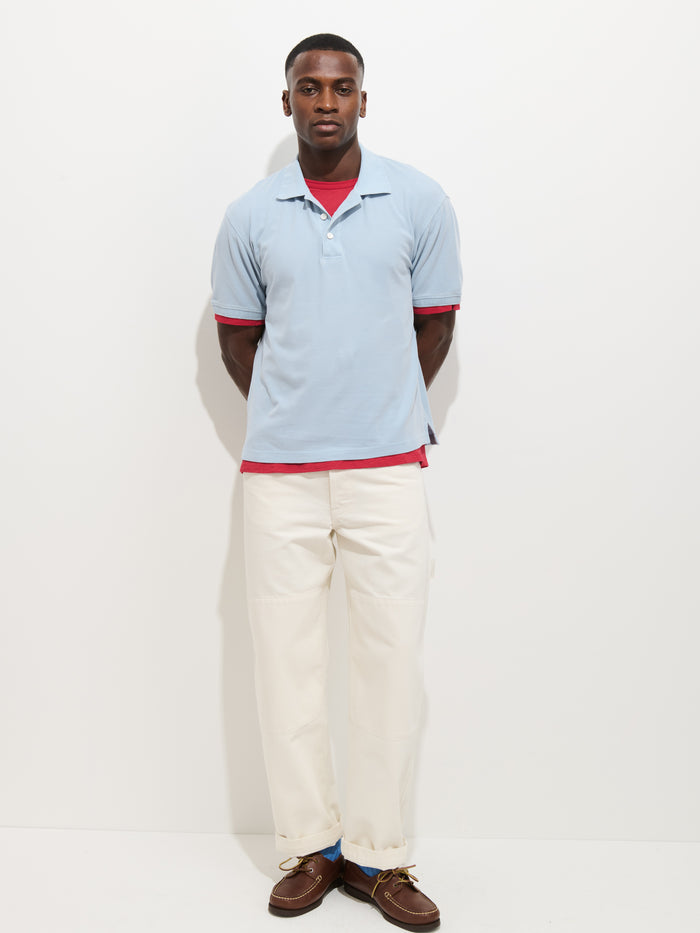 A man faces forward with a neutral expression, hands behind his back, wearing a Garment Dyed Polo In Pique Cotton (light blue) over a red T-shirt, white pants, and brown shoes against a white background.
