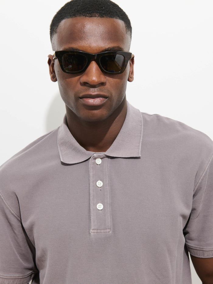 A man wearing dark sunglasses and a Garment Dyed Polo In Pique Cotton stands against a plain white background, looking directly at the camera.