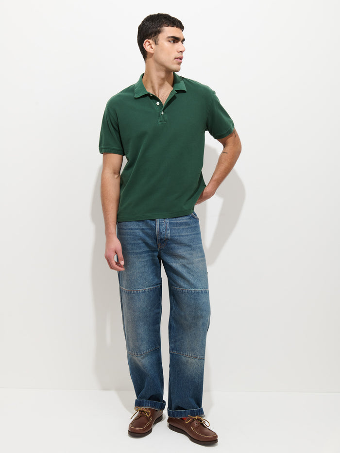 A person stands against a white background, wearing the Garment Dyed Polo In Pique Cotton in dark green, paired with blue jeans featuring visible seams and brown leather shoes. One hand is in their pocket as they look to the side.