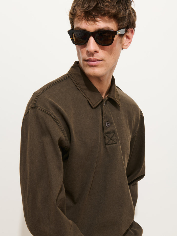 A man wearing dark sunglasses and a dark olive Garment Dyed Rugby Shirt In Cotton Jersey with a boxy fit looks to the side against a plain white background.
