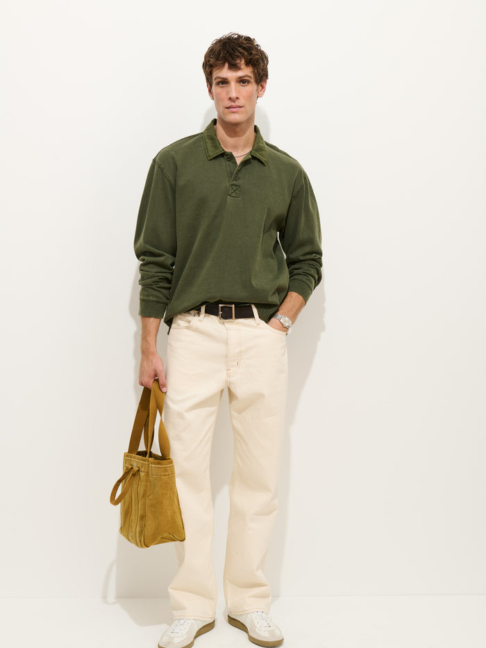 A man stands against a white background, wearing the Garment Dyed Rugby Shirt in Cotton Jersey in green with a boxy fit, cream pants, white sneakers, and holding a tan suede bag.