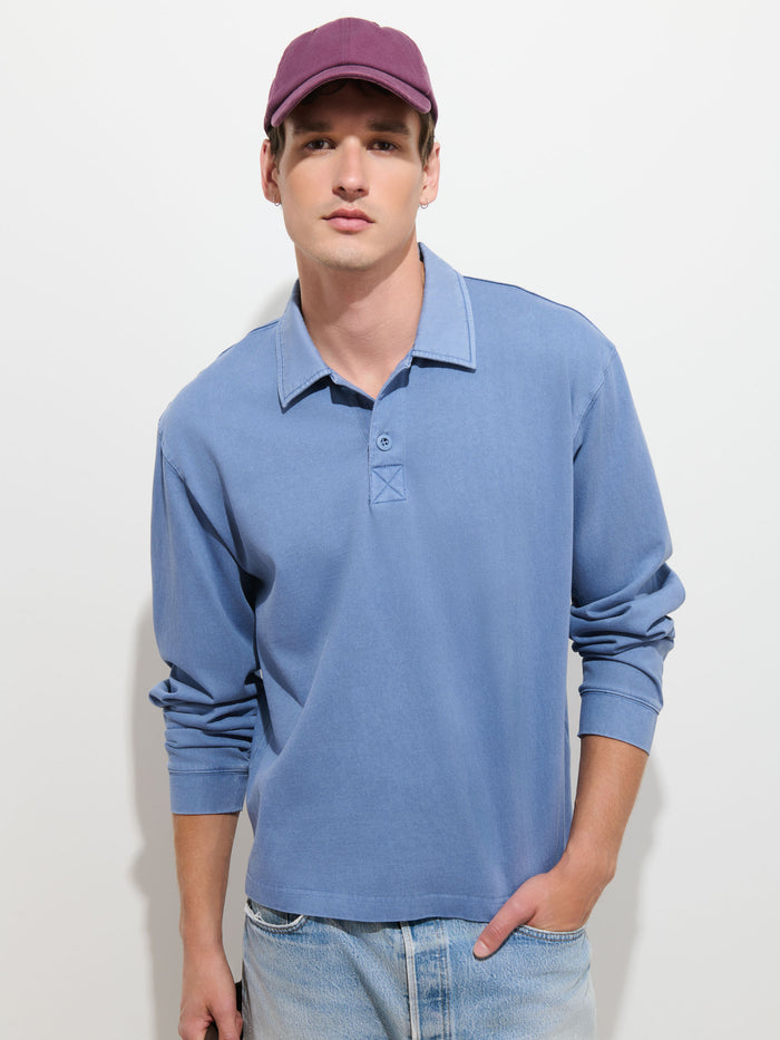 A young man wearing a purple cap, Garment Dyed Rugby Shirt In Cotton Jersey with a boxy fit, and light-wash jeans stands against a plain white background with one hand in his pocket.