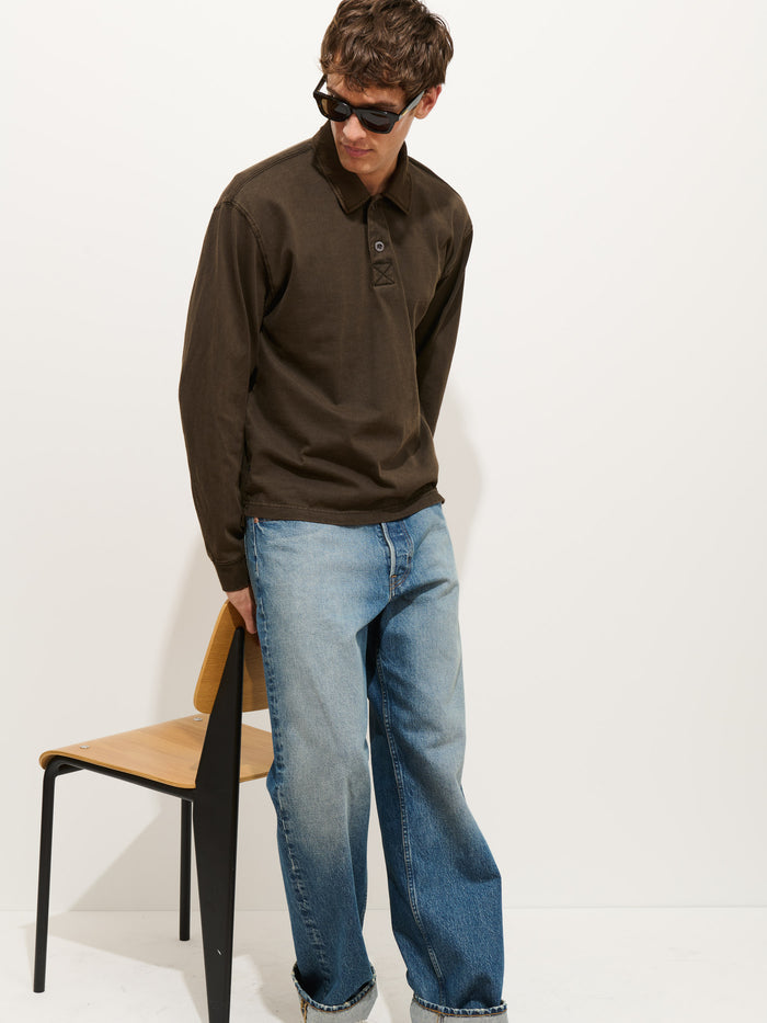 A person wearing sunglasses, a dark brown Garment Dyed Rugby Shirt In Cotton Jersey with a boxy fit, and loose-fitting blue jeans stands beside a wooden chair against a plain white background.