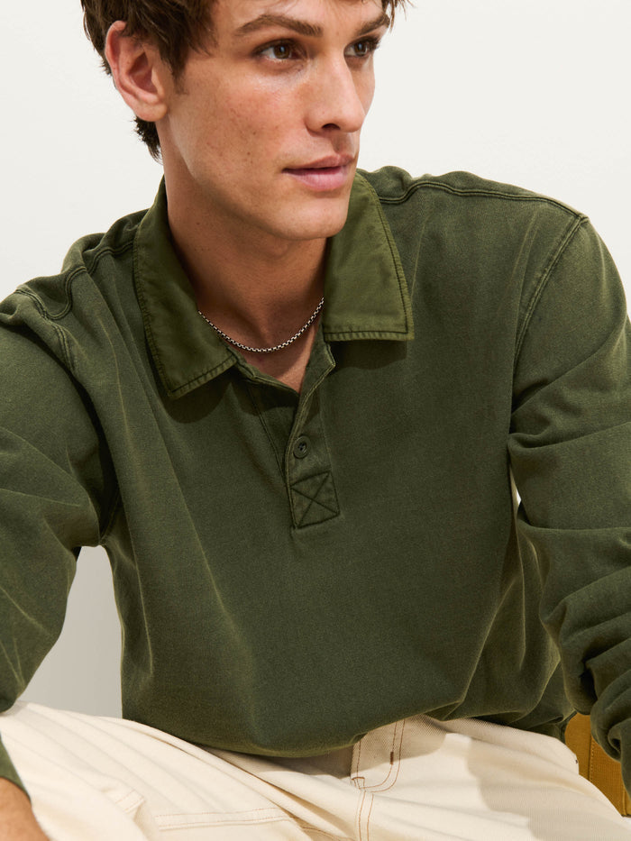 A man dressed in the Garment Dyed Rugby Shirt In Cotton Jersey, an olive green collared top, pairs it with a silver chain as he looks to the side. He sits against a light background with his hands resting on his knees.