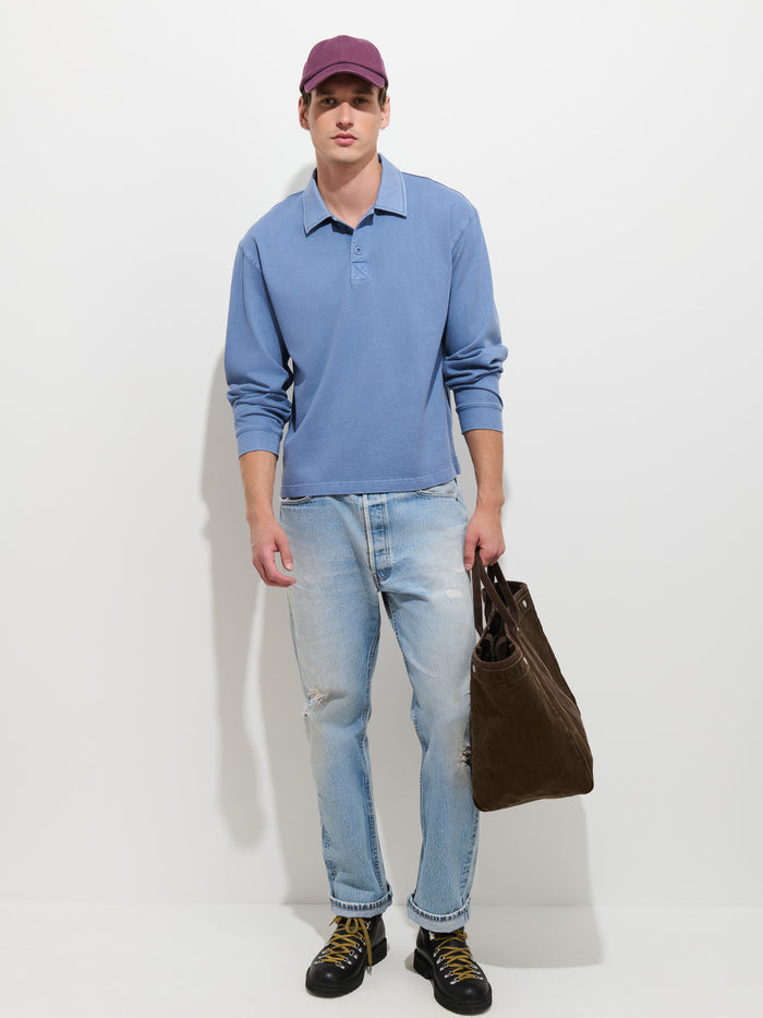 Wearing the Garment Dyed Rugby Shirt In Cotton Jersey—a blue, boxy fit piece—a person styled in a purple cap, light ripped jeans, and black shoes holds a brown bag while standing against a plain white wall.