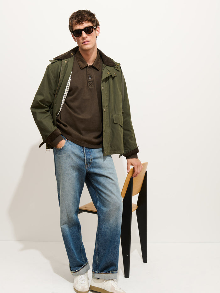 A person wearing sunglasses, a green Garment Dyed Rugby Shirt In Cotton Jersey with a boxy fit, brown polo shirt, blue jeans, and white sneakers leans back on a modern wooden chair against a plain white background.