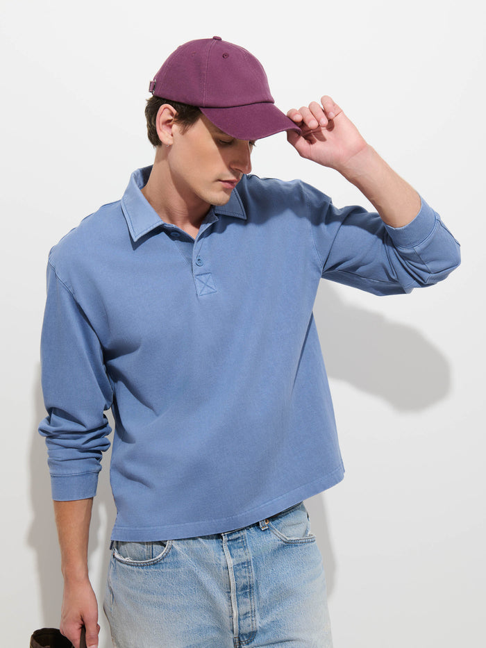 Wearing the Garment Dyed Rugby Shirt in Cotton Jersey with light denim jeans, a person tilts a purple cap while looking down, posed against a plain white background.
