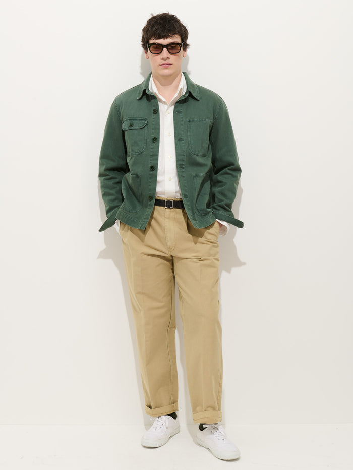 A man in dark sunglasses, a white shirt, beige pants, and white sneakers wears the Garment Dyed Work Jacket In Recycled Denim, standing with hands in his pockets against a plain white background.