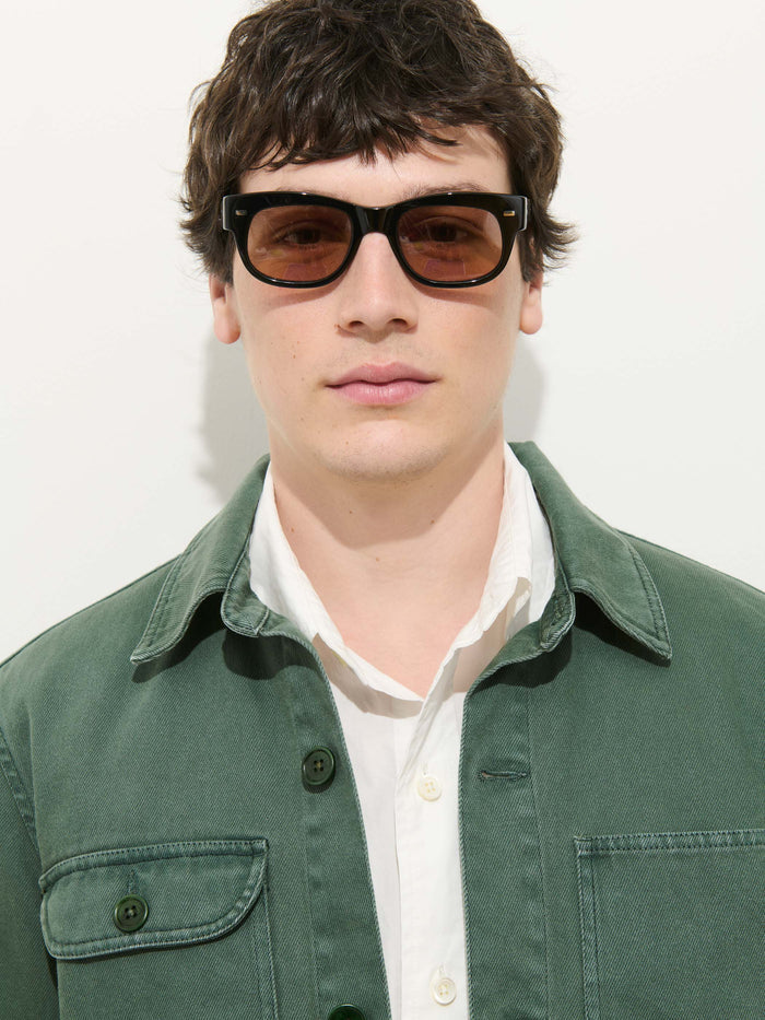 A young man with short dark hair wears black sunglasses, a white button-up shirt, and the Garment Dyed Work Jacket In Recycled Denim, standing against a plain white background.