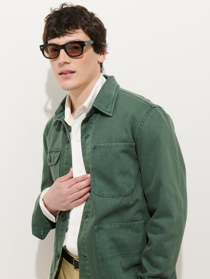 A person with short dark hair and sunglasses wears the Garment Dyed Work Jacket In Recycled Denim over a white button-up shirt and beige pants, standing against a plain white background with one hand on their chest.