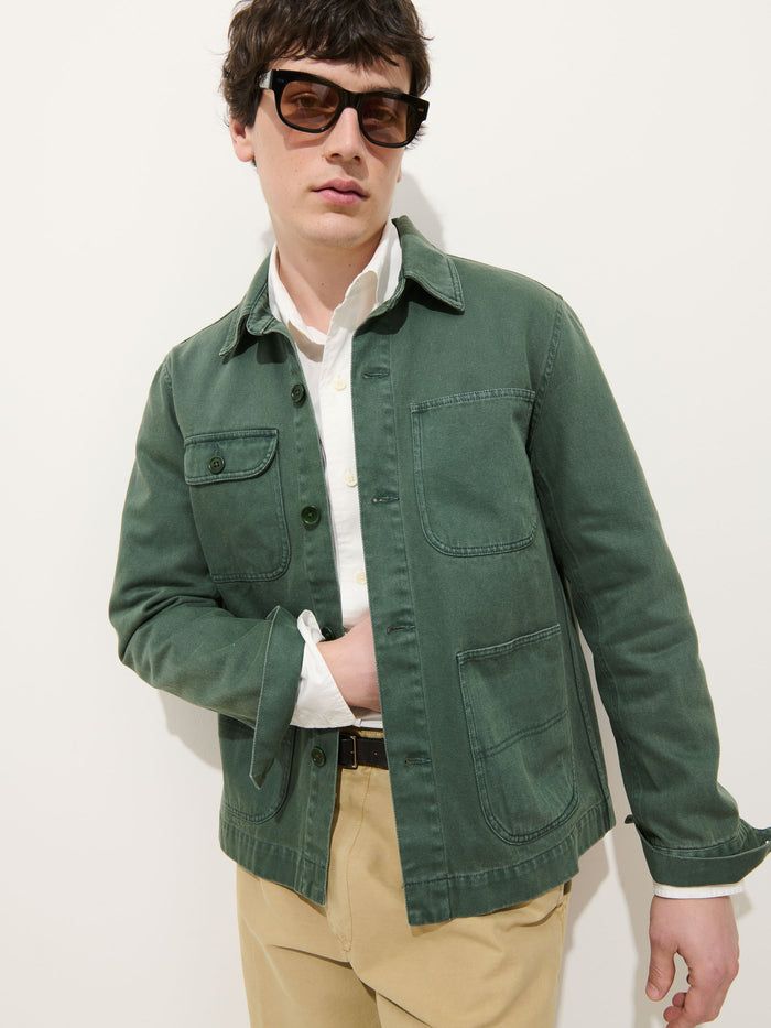 A man in large dark sunglasses, a white button-up shirt, the Garment Dyed Work Jacket In Recycled Denim, and beige pants stands against a plain white background.