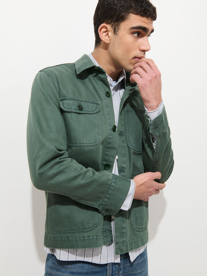 A young man with short dark hair wears the Garment Dyed Work Jacket in Recycled Denim over a light striped shirt and blue jeans, standing against a white background, looking to the side with his hand on his chin.